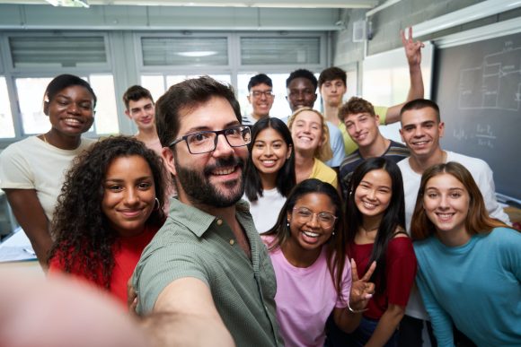 Happy selfie of young group of students taking a photo with their male teacher in the classroom, celebrating the end of course. Classmates from different countries, looking at camera with big smiles.